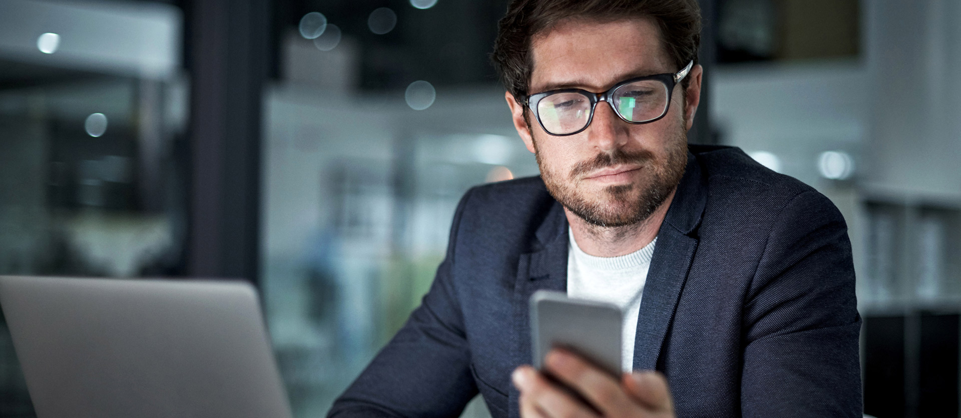 Mann sitzt vor seinem Laptop und sieht auf sein Smartphone