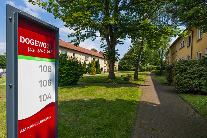 Wegweiserschild der DOGEWO21 mit Hausnummer und Häusern im Hintergrund