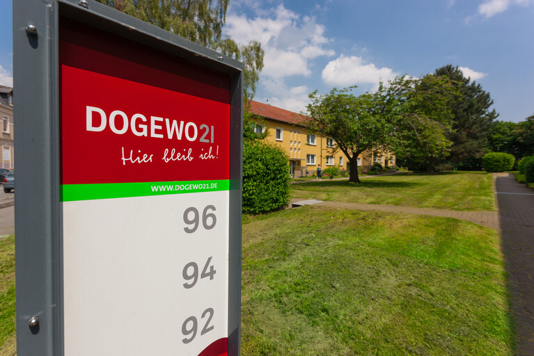 Wegweiserschild der DOGEWO21 mit Hausnummer und Häusern im Hintergrund